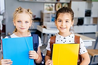 To skolejenter med ryggsekker som holder en blå og en gul mappe i et klasserom