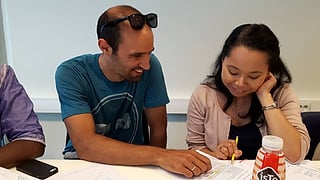 En mann og en kvinne lener seg over arbeidsark ved et skrivebord, kvinnen holder en gul penn