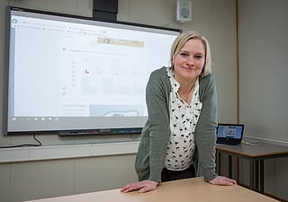 Smilende kvinne som lener seg på et skrivebord foran en stor skjerm og viser et digitalt matematikkarbeidsark