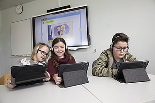 Tre elever med hodetelefoner bruker nettbrett ved et skrivebord, en stor skjerm viser en læringsapp bak dem