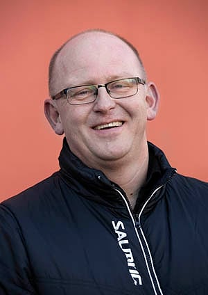 Smilende skallet mann med briller iført en svart Salming-jakke mot en oransje bakgrunn