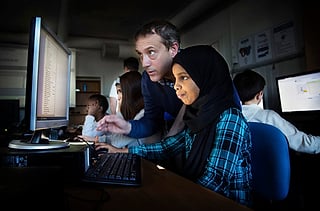 Lærer lener seg over for å hjelpe en jente i hijab ved en datamaskin i et klasserom